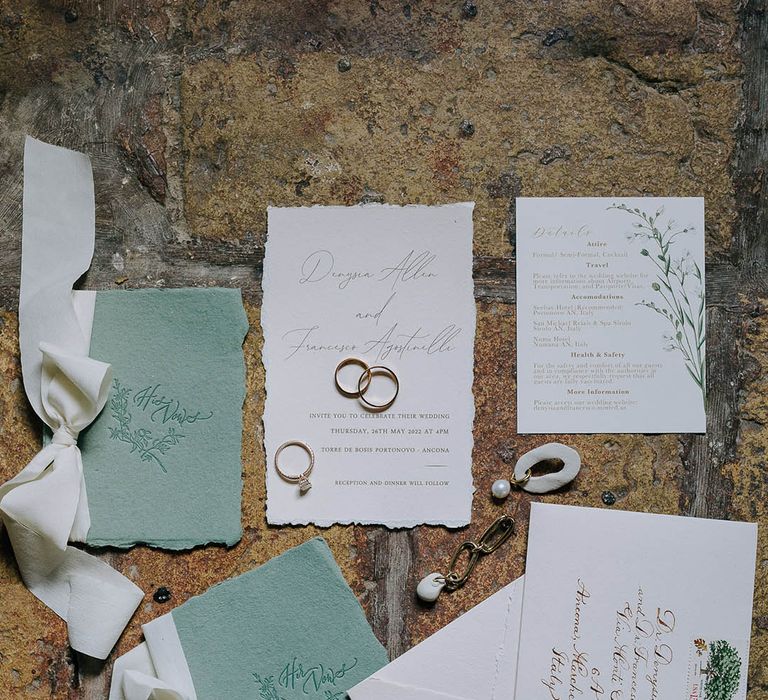 Sage green vow books tied with a white ribbon bow accompanied by white rustic wedding invitations, wedding rings, engagement ring and other jewellery 