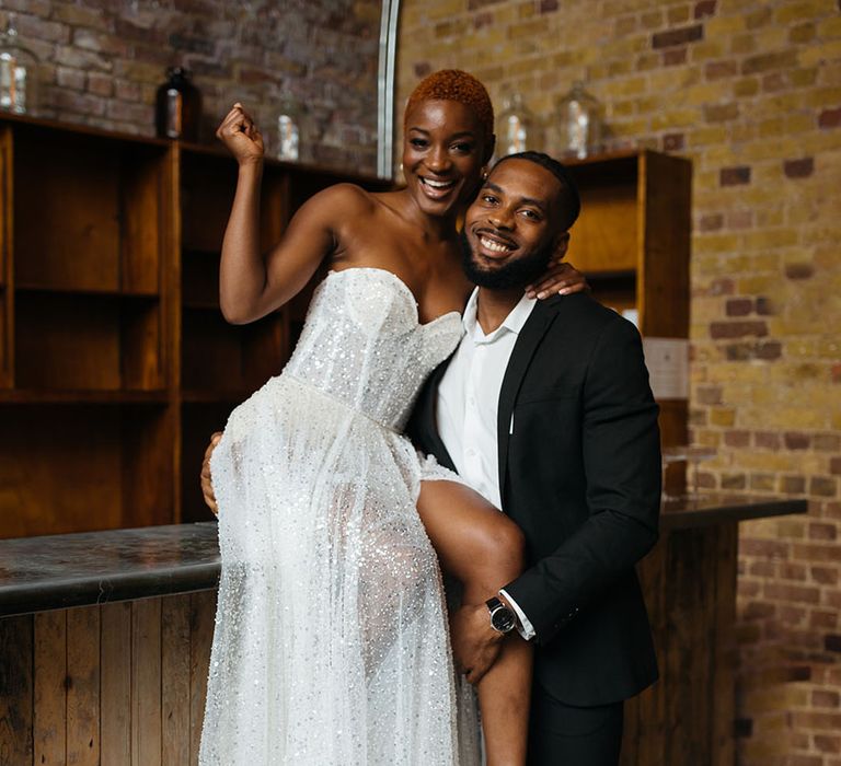 Bride sitting on the bar wearing sparkly sweetheart strapless dress and leg slit and groom embracing her black Topman tux