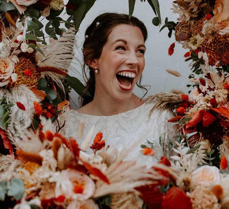 Bride in long sleeve lace vintage wedding dress with pearl earrings and pearl hair clips standing in large autumnal floral arch with roses, peonies, foliage and pampas grass