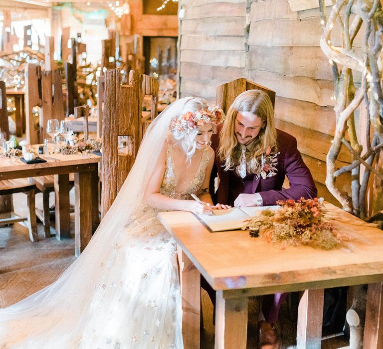 Bride in flower crown and veil sits beside her groom in maroon suit to sign marriage certificate 