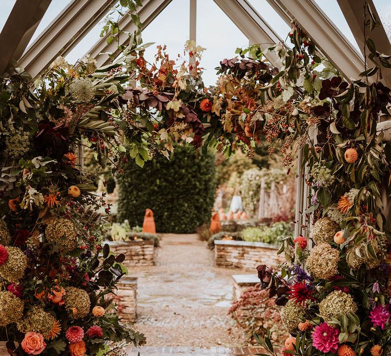 Large luxe floral arch in glasshouse wedding venue with white and pink carnations, garden roses, red strawflower, eucalyptus, baby’s-breath and dried flowers 