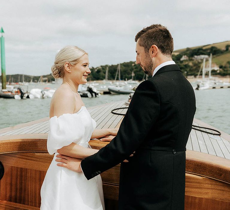 Bride in puffed sleeve Jesus Peiro wedding dress stands beside her groom during boat road for Salcombe wedding 