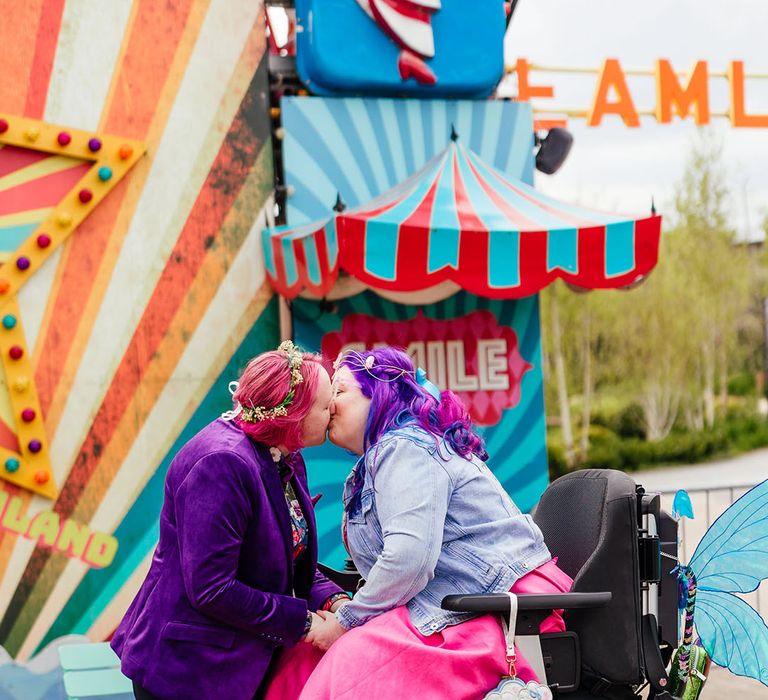 Bride in personalised denim jacket and bright pink wedding dress with blue tights in a wheelchair with sparkly blue fairy wings kissing bride in purple velvet blazer and black suit trousers with purple bowtie, pink patterned pocket square and colourful patterned shirt