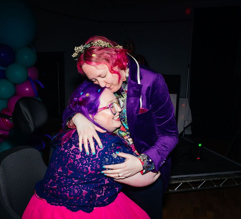 Bride in purple velvet blazer and black suit trousers with purple bowtie, pink patterned pocket square and colourful patterned shirt embracing bride wearing personalised denim jacket, pink wedding dress and blue tights at wedding party 