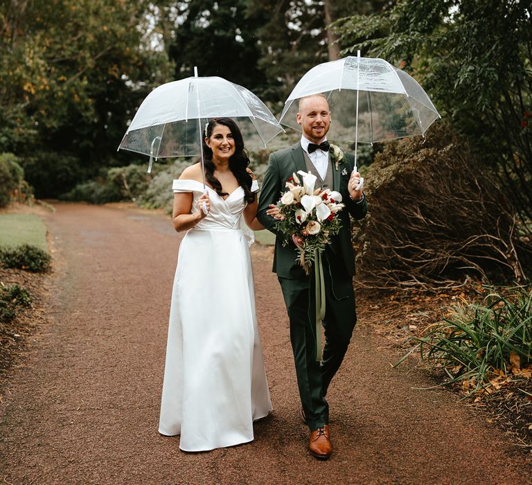 Bride in off-the-shoulder wedding dress walks alongside her groom in green suit beneath clear umbrellas 