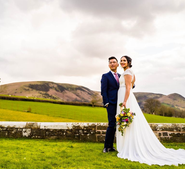 Bride in open back lace wedding dress embraces her groom outdoors surrounded by lush rolling hills 