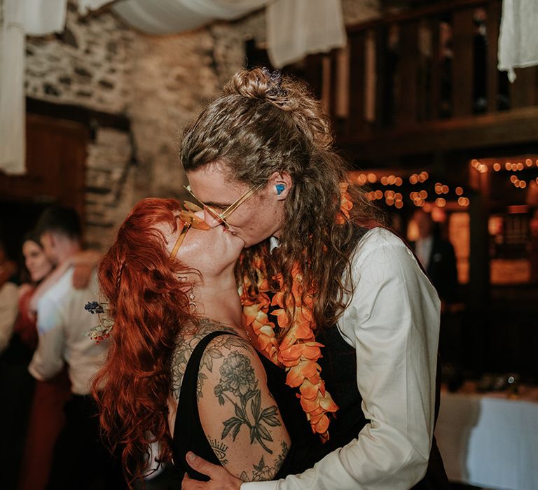 Groom in checkered waistcoat, white shirt and orange lei kissing bride in black wedding dress and orange wedding sunglasses 