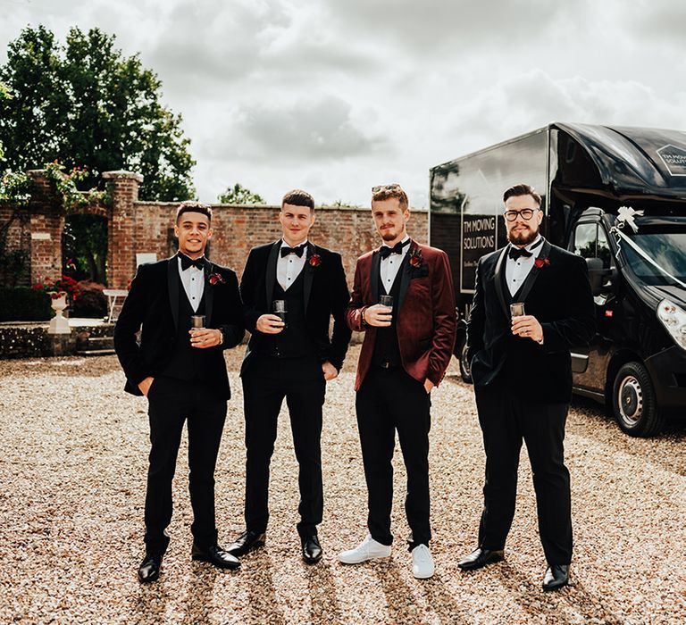 Groomsmen in black tuxedos with the groom in a burgundy velvet tuxedo next to their black van wedding transport decorated with white ribbon 