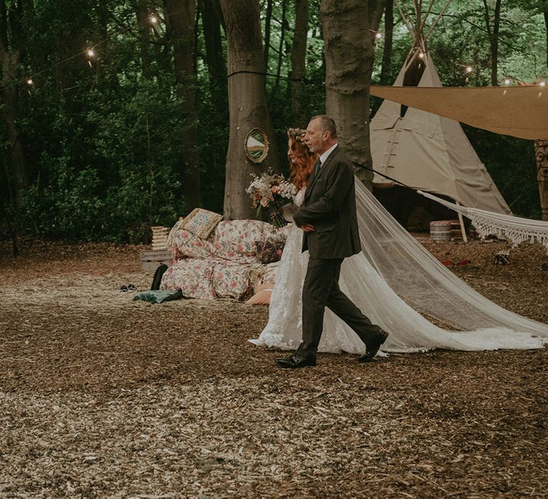 Father of the bride in a tweed suit walks the bride in lace wedding dress down the aisle at outdoor wedding 