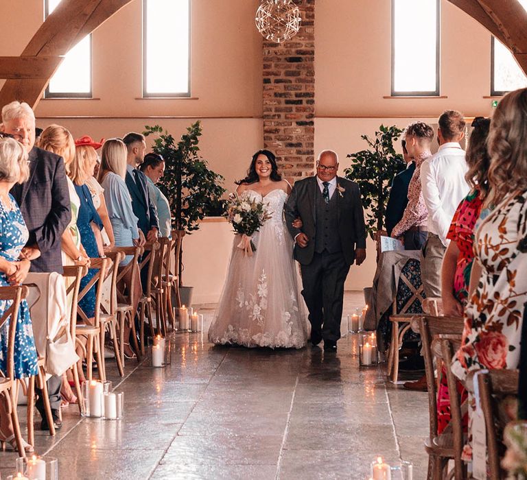 Bride in off the shoulder lace wedding dress walking down the aisle decorated with white pillar candles with her father in a grey suit 