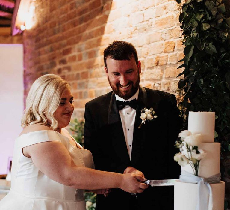 Bride wearing satin off the shoulder wedding dress cutting three tiered wedding cake with groom in classic black grooms three piece tuxedo with white rose boutonniere and black bowtie 