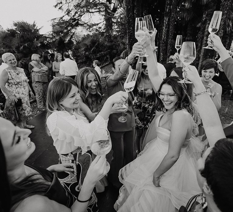 bride with glasses wearing a strapless wedding dress raising her glass surrounded by wedding guests