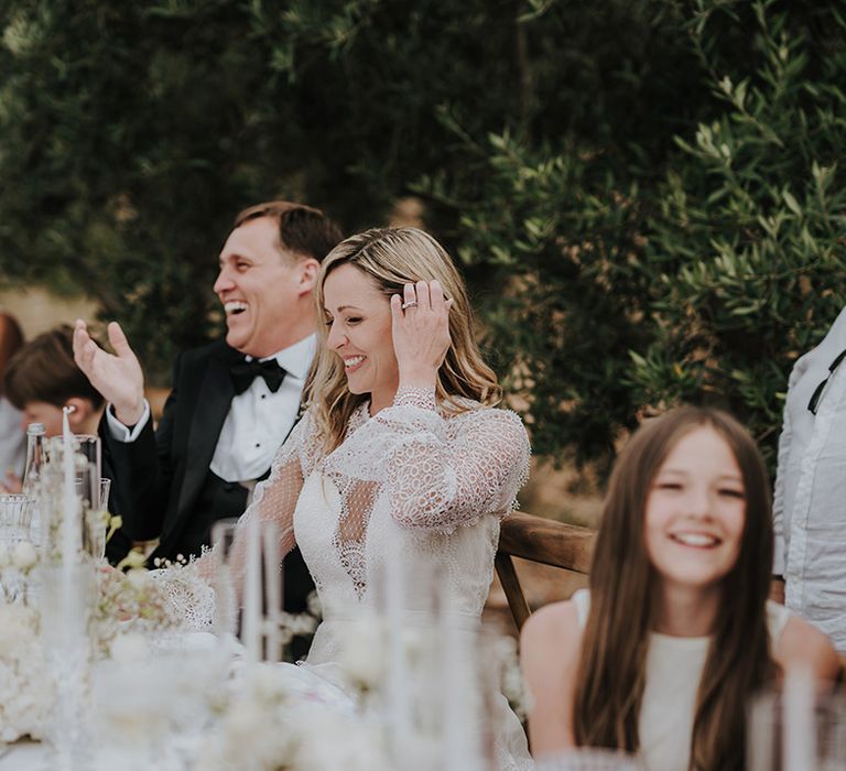 bride in a long sleeve lace wedding dress and groom in a tuxedo laughing during the wedding speeches at outdoor pure house ibiza wedding reception