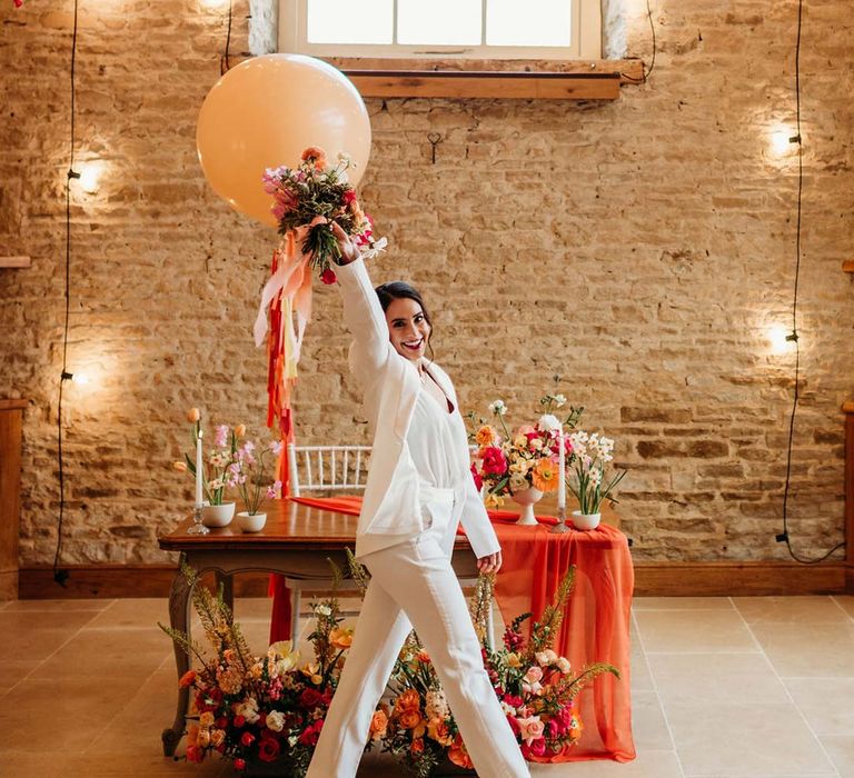 Bride in white bridal suit with closed toe bright green wedding heels holding large wedding balloon decoration in the reception room of Merriscourt wedding venue with reception room with metallic pink, white, orange and yellow balloons, wedding fairy lights decorations and wooden table with orange table runner and mixed seasonal flower arrangements 