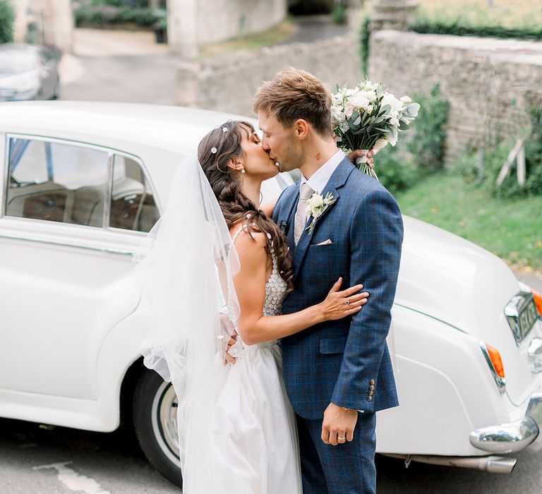 groom in a navy check suit kissing his bride in a fitted wedding dress in from of their classic white wedding car 