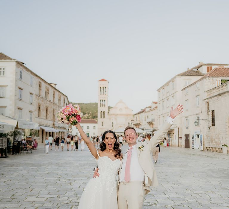 Bride in 3D lace off-the-shoulder wedding dress holds up wedding bouquet with groom in linen suit and pink tie in Old Town Hvar at destination wedding in Croatia