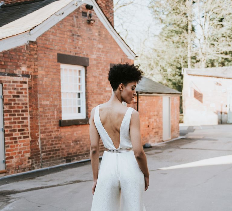 A bride stands in the street in a low back jumpsuit. Her hair is short to the back and sides and she has curls piled on top of her head.