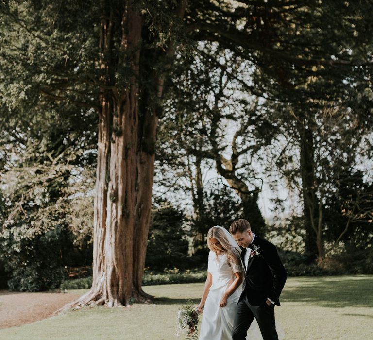 Bride in white Charlie Brear dress, white lace Augusta Jones top and full length veil walking with groom in black velvet Hugo Boss suit in the grounds at Caswell House wedding