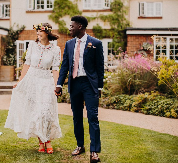 Groom in a navy suit and pink tie holding hands with his bride in a white Broderie Anglaise wedding dress as they arrive for their outdoor wedding reception