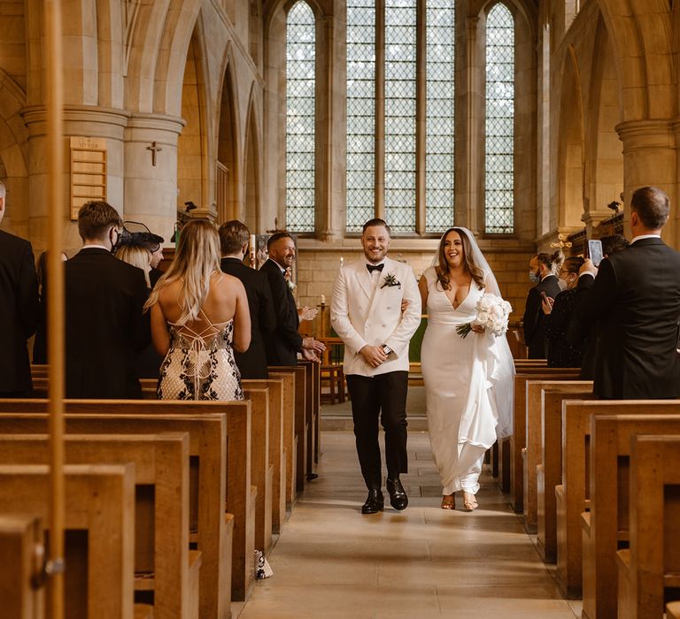 Smiling groom in white tuxedo jacket walks back down the aisle with smiling bride in white Made With Love wedding dress and white chapel length veil