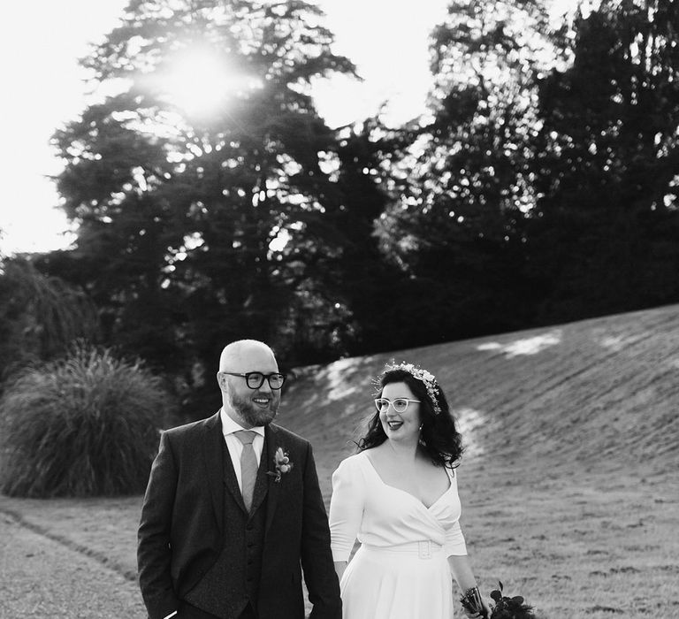 Groom in navy suit and yellow tie walks through gardens with bride in white cat eye glasses and bridal headband holding white and pink rose and pampas grass bouquet