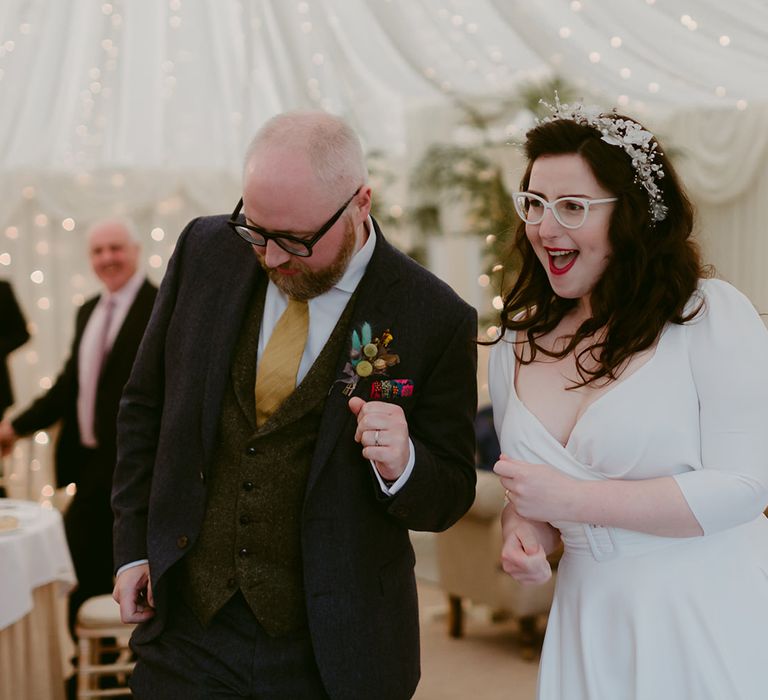 Bride in white Rime Arodaky wedding dress with bridal headband and white cat eye glasses dances with groom in navy suit and yellow tie