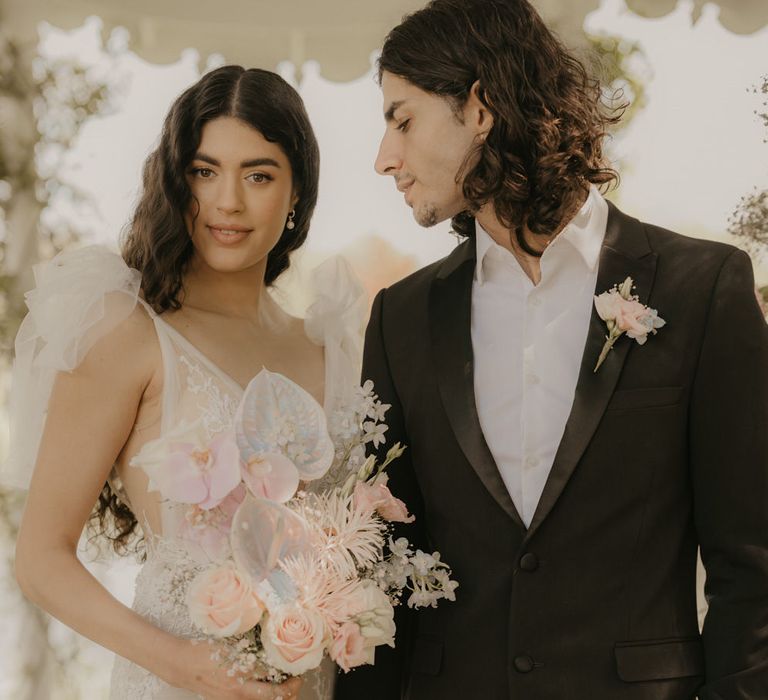 Groom in a white shirt and black blazer looking at his bride in a romantic lace wedding dress holding a pastel wedding theme bouquet 