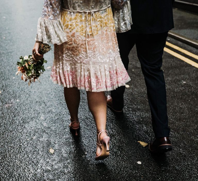 Bride and groom walking down the street, bride wears pastel embellished dress and holds wedding bouquet at her side