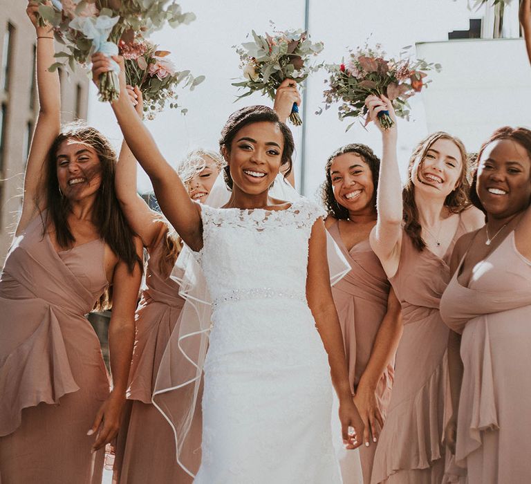 Bride in lace wedding dress with her bridesmaids in pink dresses holding their bouquets in the air 