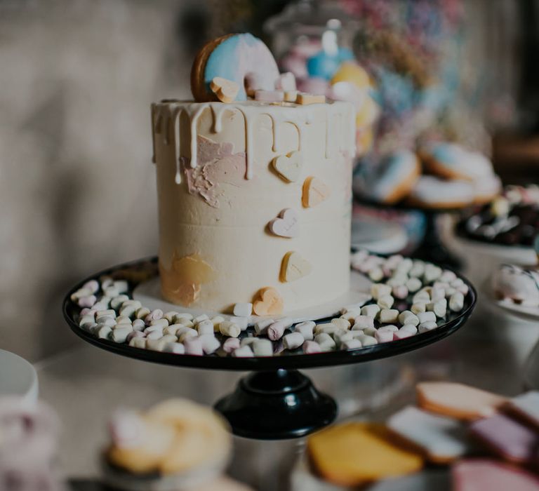 Pastel buttercream, drip wedding cake decorated with mini doughnuts, sweets and marshmallows on a black cake stand 