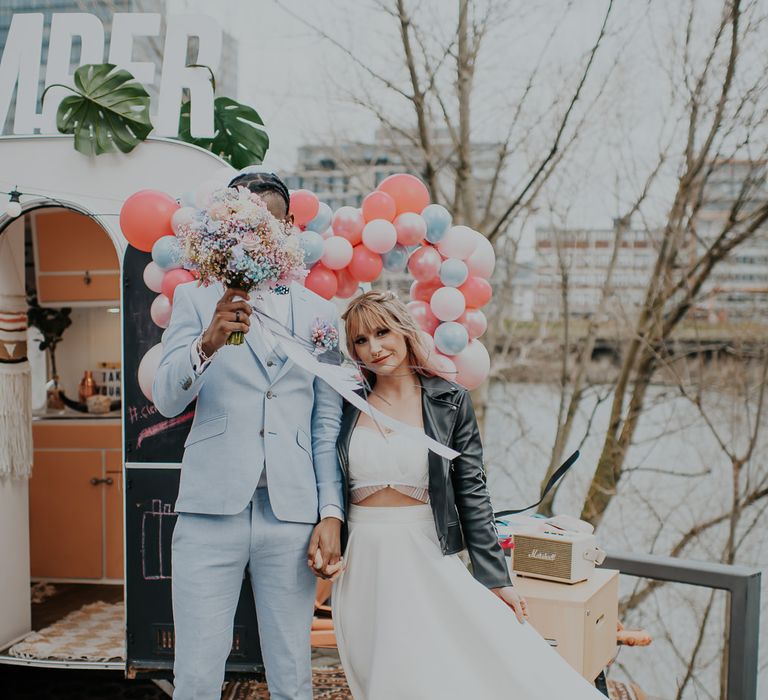 Bride in separates and leather jacket standing with her groom in a pale blue wedding suit outside a street van