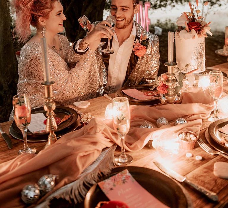 Bride with pink curled hair and gold sequinned dress raises tropical print Champagne flute with groom in silver sequinned suit jacket sat at table outside at festival themed wedding