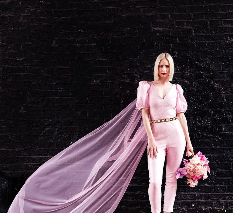 Stylish bride in an alternative wedding dress pink jumpsuit with cape, pink shoes and blush pink bouquet 