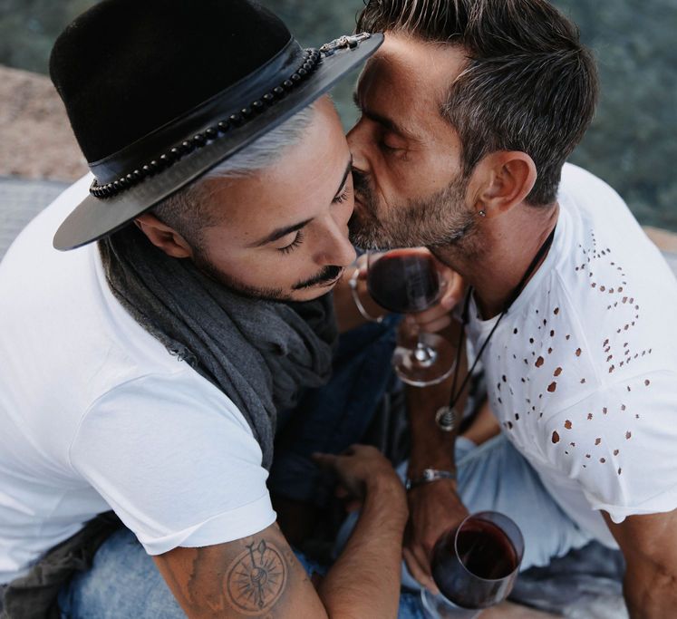 A gay couple kiss during engagement shoot. They both hold a glass of red wine and one wears a hat. Photography by Stephanie Shenton Photography. 