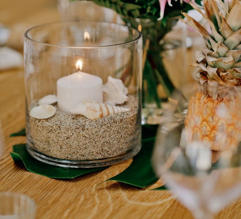 Candles in holders filled with sand and shells surrounded by vibrant flowers