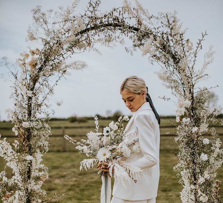 Stylish bride in a wide leg trouser suit holding an all white flower wedding bouquet under a flower arch 