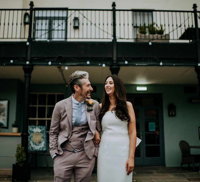 Bride in white strapless Rebecca Vallance Dress stands arm in arm with groom in light brown Moss Bros suit and blue tie outside at the Hotel du Vin Harrogate