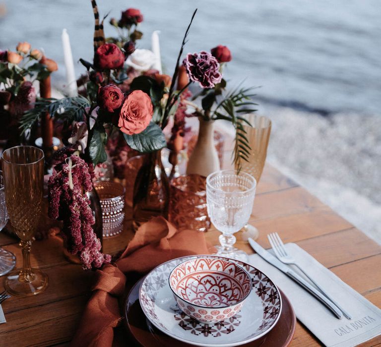 Terracotta toned tableware with floral centrepiece containing feathers for beachside ceremony in Ibiza