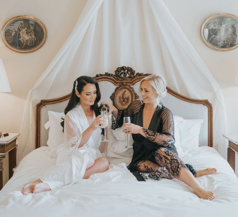 Bride with long dark curled wedding hair and long white bridal dressing gown sits on bed with maid of honour wearing black lace dressing gown as they both raise a glass of sparkling wine