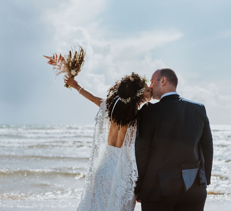 Bride lifts her dried floral bouquet in the air in celebration on the beach as she leans in to her husband