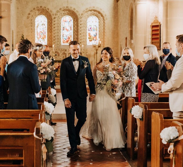 Bride in white cami wedding dress holding multi-coloured bridal bouquet and groom in blue suit and bow tie smile as wedding guests throw confetti after church wedding ceremony