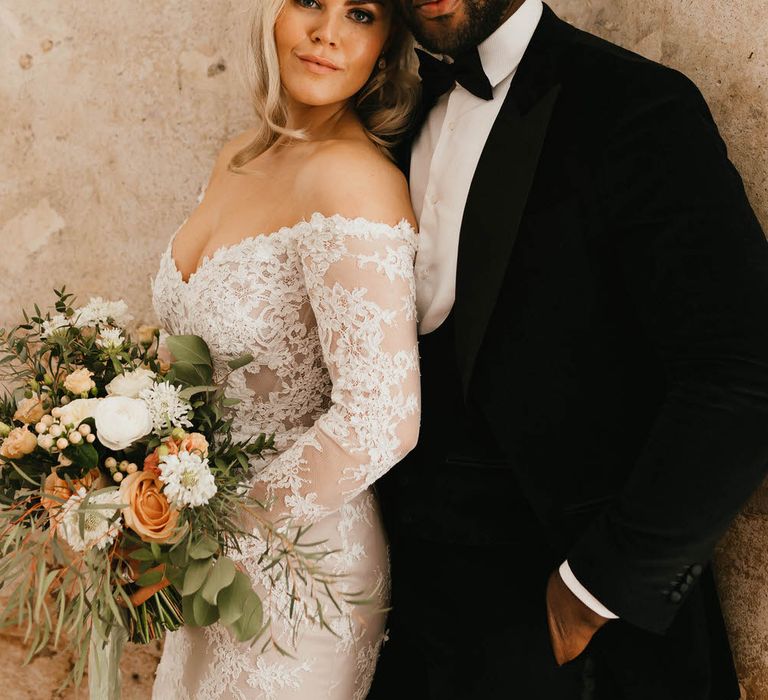 Bride and groom against plain industrial style wall, leaning on each other. The bride is holding her bouquet.