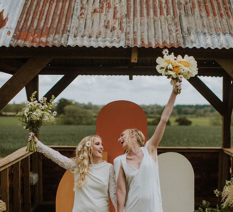 two brides raising their wildflower bouquets in the air standing at the retro inspired altar 