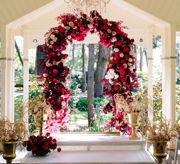 Bright red and pink floral arch stands at the end of the aisle which is lined with white chairs