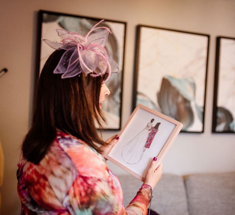 Wedding guest in colourful long sleeved dress and pink fascinator holds framed illustration of herself and the bride at The West Mill Derby