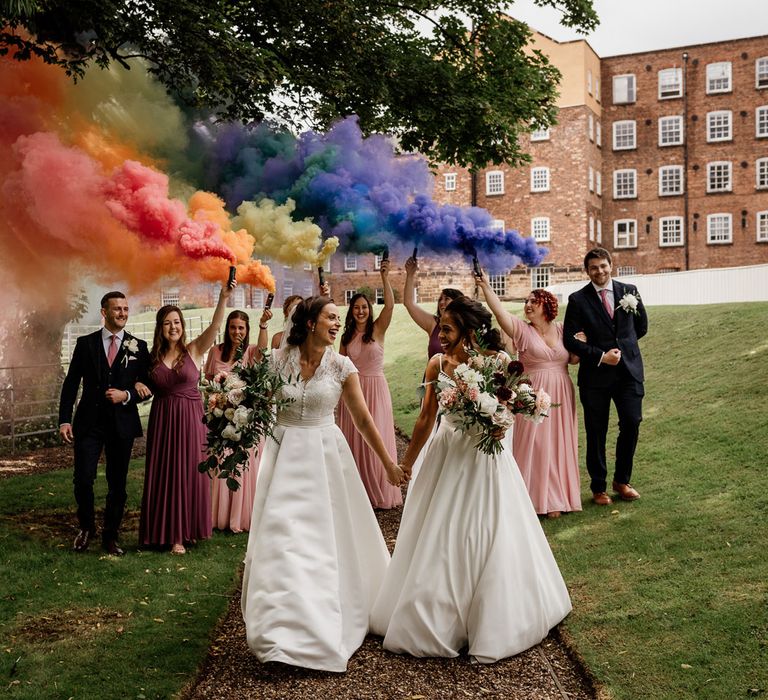 Bride in white cami wedding dress holds hands with bride in white lace capped sleeve wedding dress as bridesmaids in pink bridesmaids dresses and groomsmen hold rainbow coloured smoke bombs behind them outside The West Mill Derby