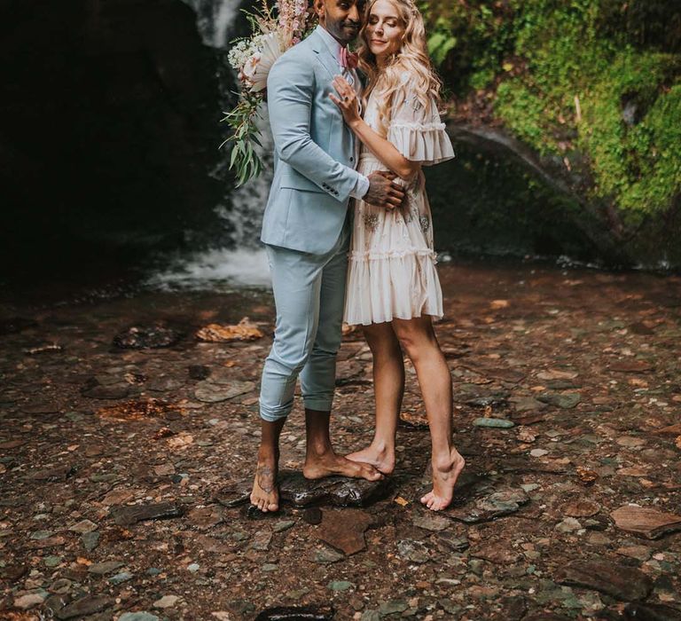 Bride & groom embrace outdoors in front of waterfall on their wedding day