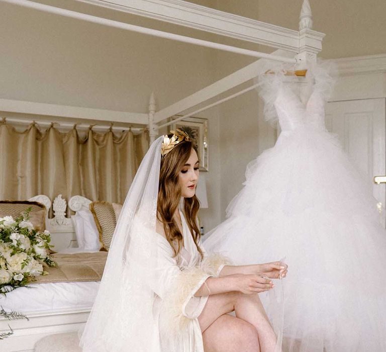 Bride on the wedding morning in a getting ready robe and lace veil putting on her embellished wedding shoes with her tulle wedding dress handing on the four poster bed 