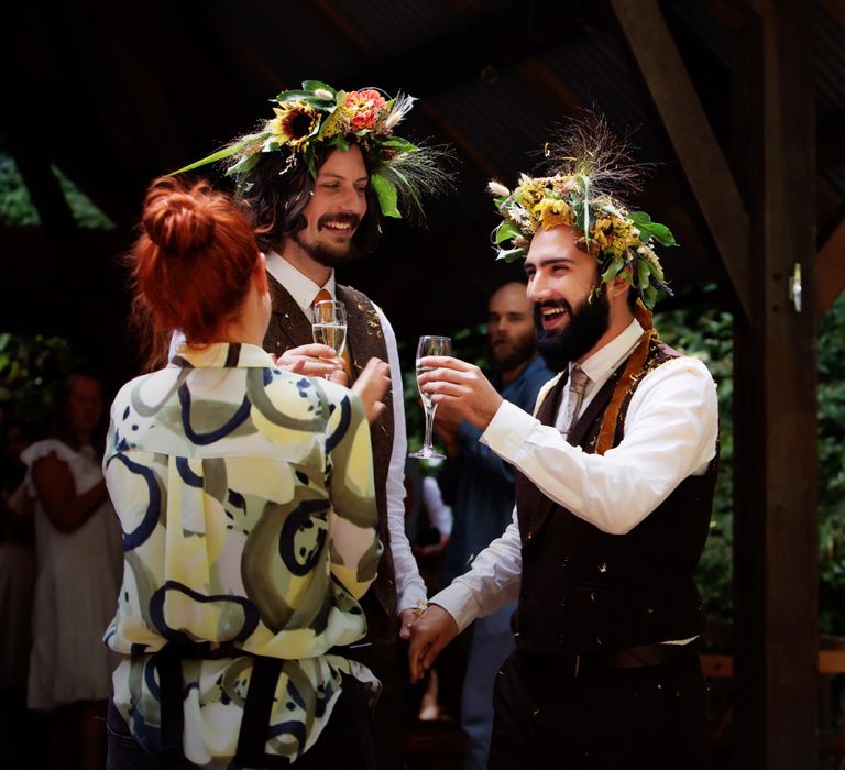 Grooms laugh and smile whilst wearing floral crowns as they lift champagne whilst covered in confetti 