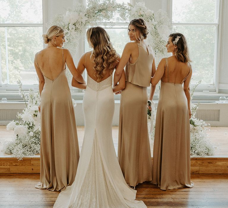 Bridesmaids in different gold satin bridesmaid dresses by Constellation Ame arm in arm with their bride in a fitted slip wedding dress